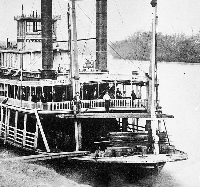 A vintage black and white photograph of a steamboat docked on shore with people on board.