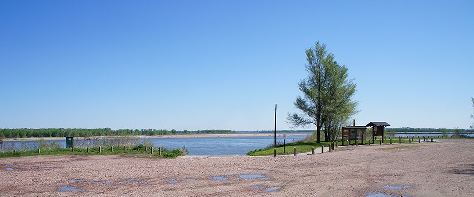 A flat gravel surface area with a single tree and utility pole stands in the scene. A river is in the background. Treeline makes the horizon.