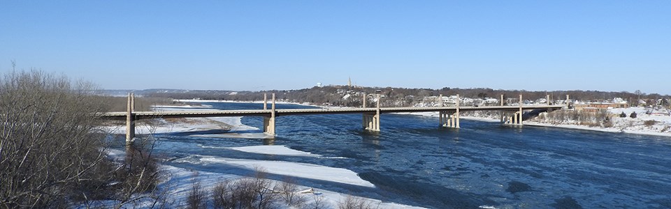 A winter landscape of snow and ice of the Missouri river with a bridge and town in the background.