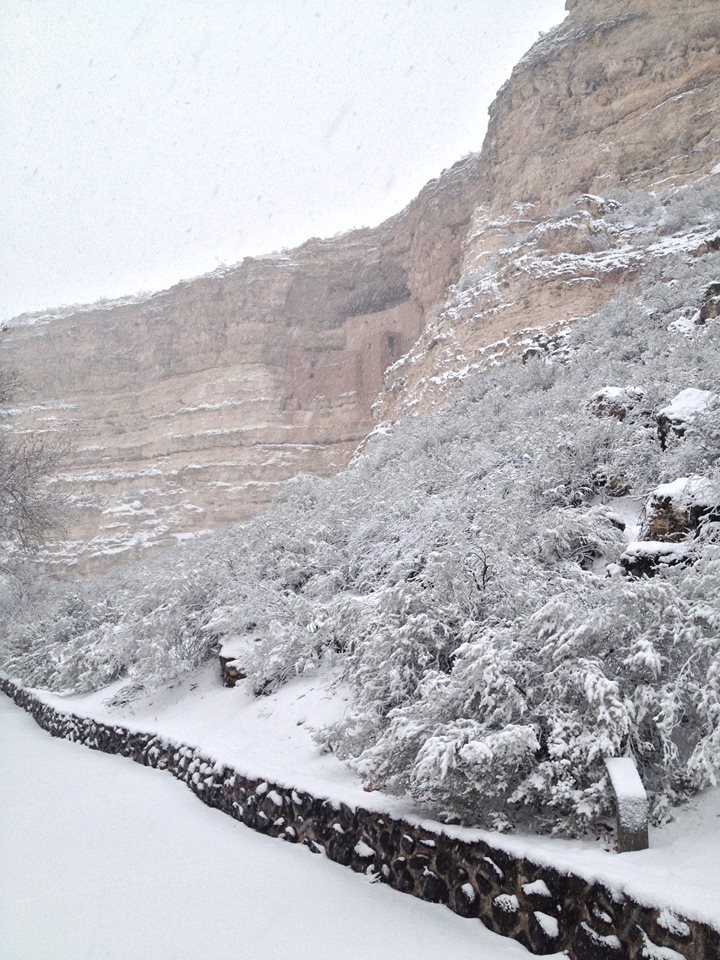 A trail with grey bushes and a cliff covered in snow, and the Montezuma Castle cliff dwelling farther down the side of the cliff.