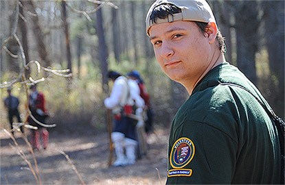 An NPS volunteer looks at the camera while living historians assemble behind him. An NPS volunteer looks at the camera while living historians assemble behind him.
