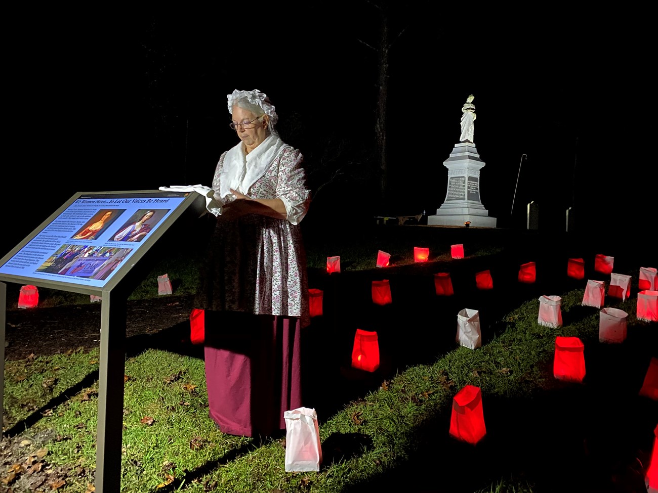 Woman standing next to wayside exhibit with red luminaries behind her at night. Monument lit up in the background.