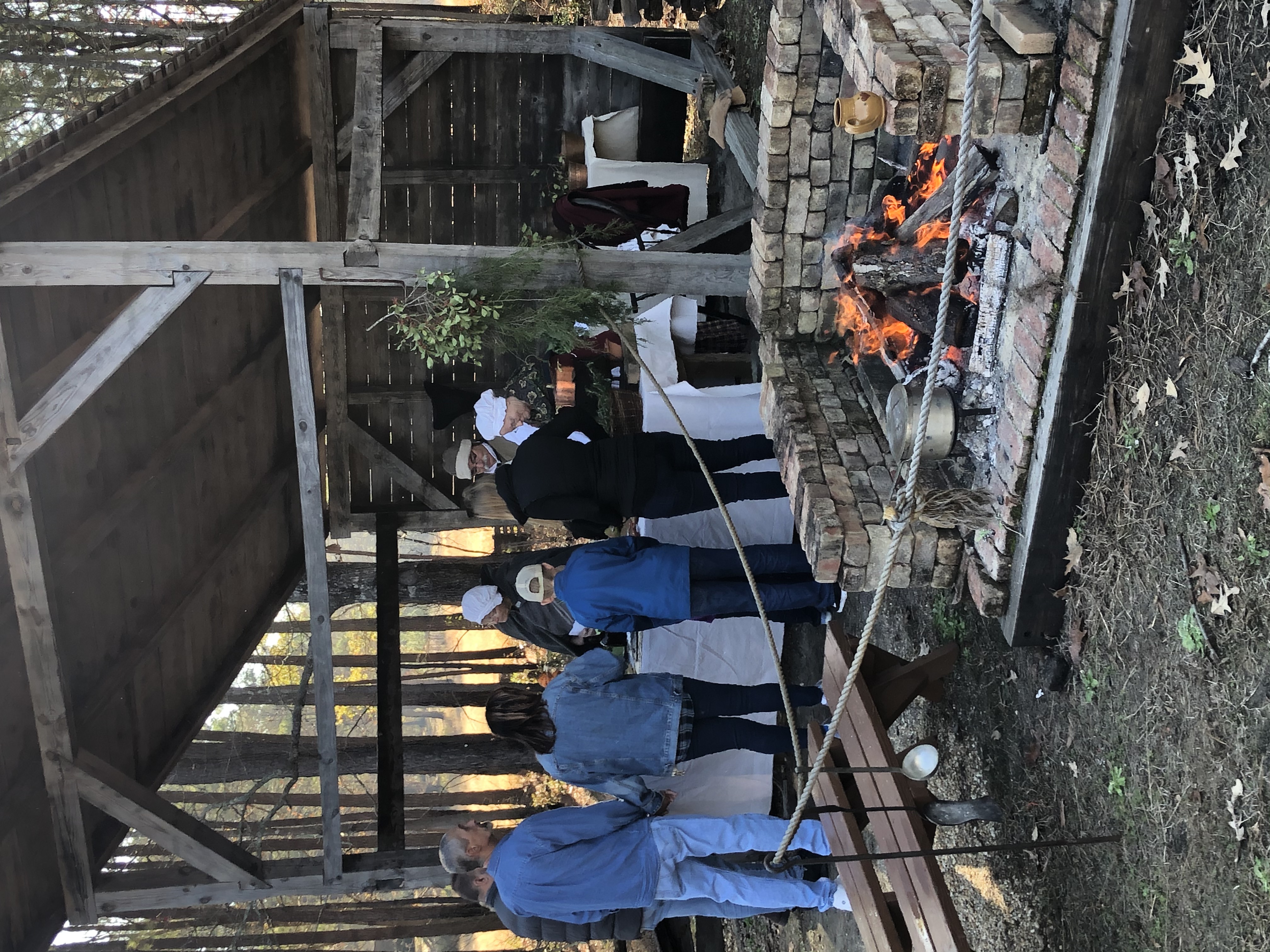 Visitors standing in front of a lean to. Campfire burning in a hearth in the foreground.