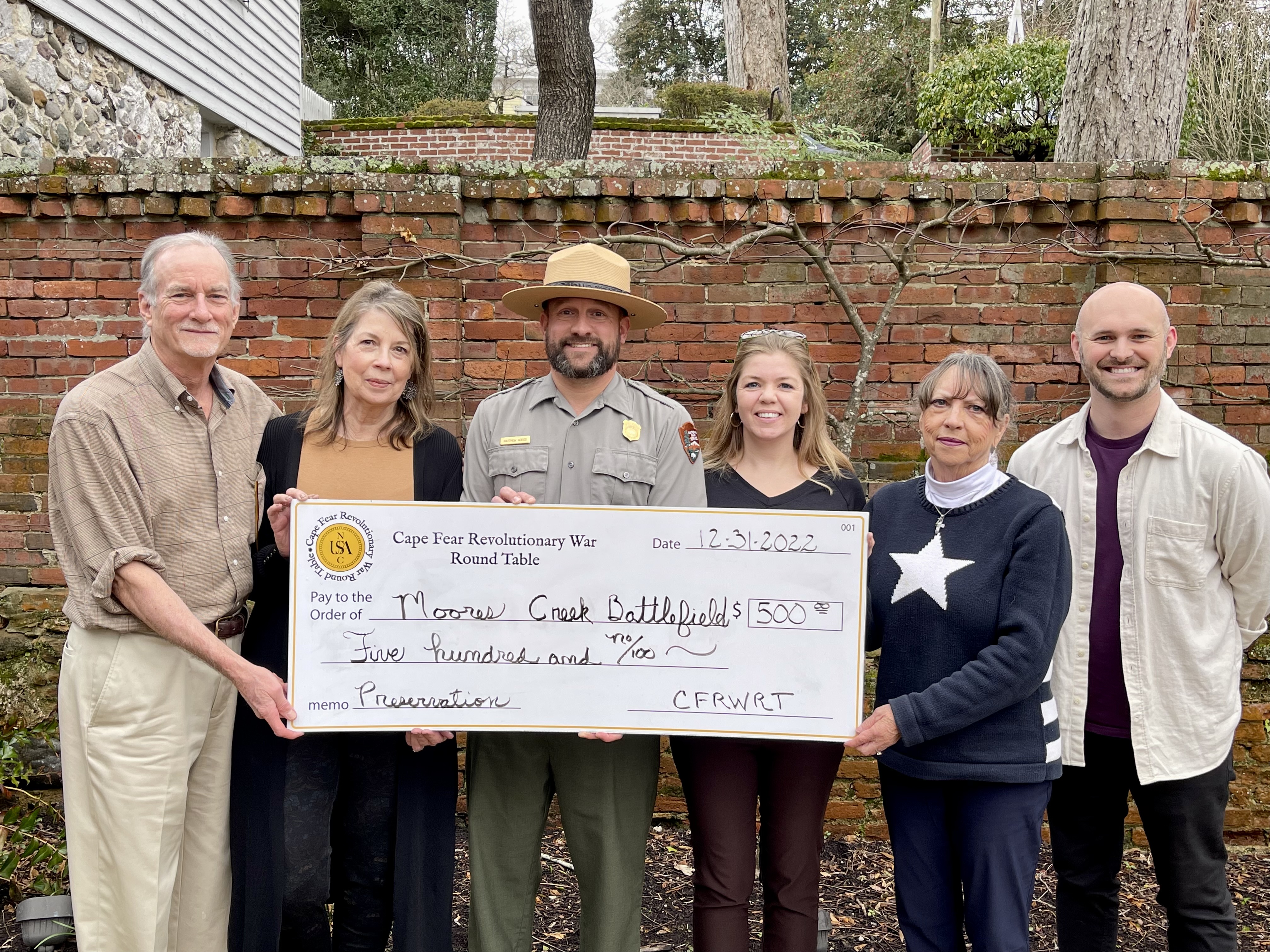 Five people standing in front of a brick wall holding a giant check for 500 dollars.