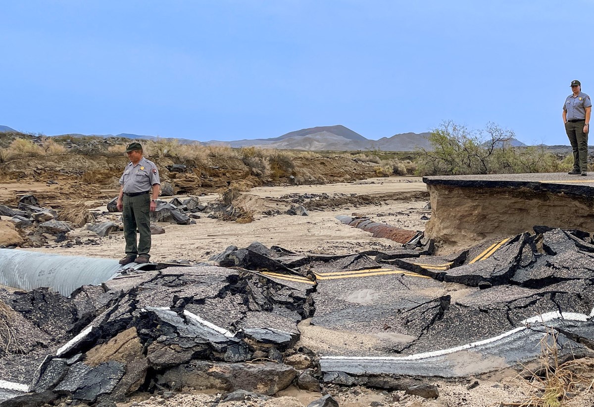 Two Park Rangers Stand on a section of washed out road and broken debris