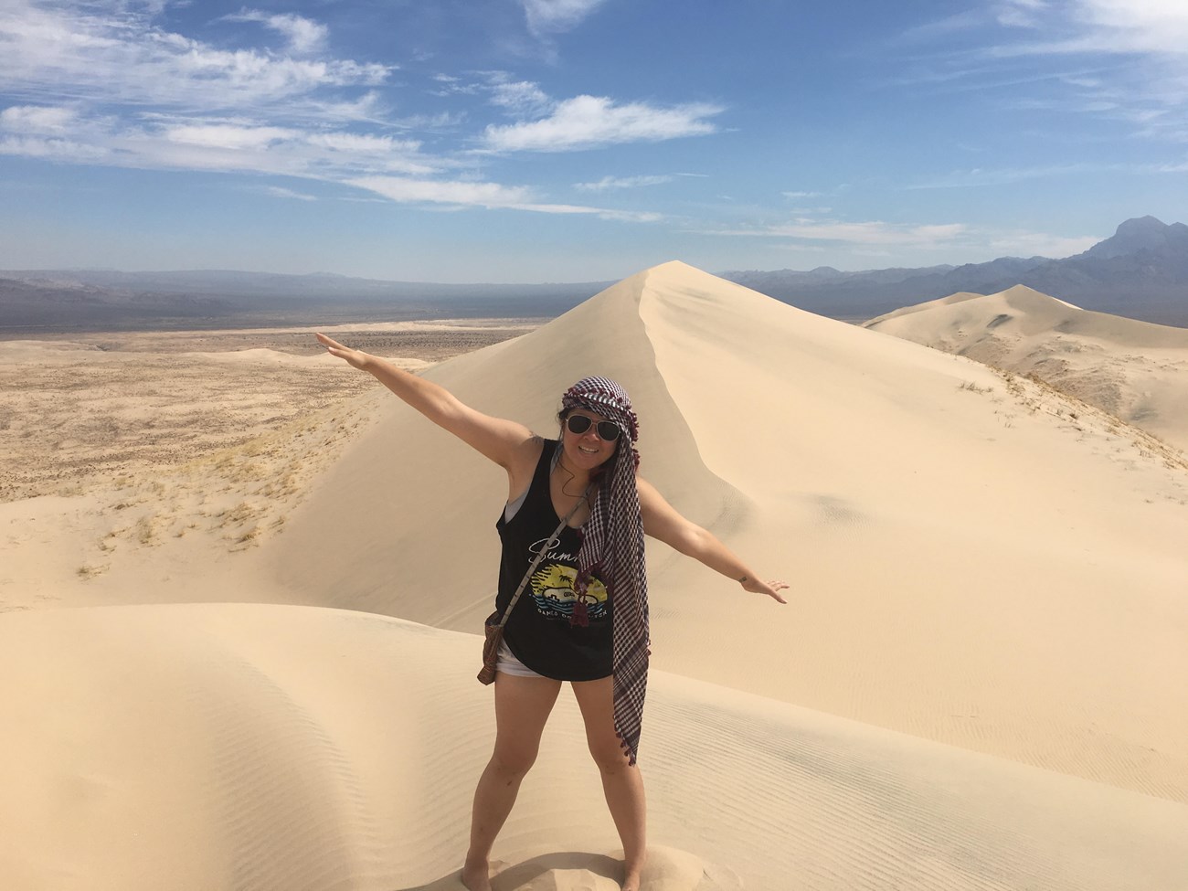 A female hiker wearing white shorts, a black tank top, and a fabric head covering smiles with arms outstretched on top of the Kelso Sand Dunes.