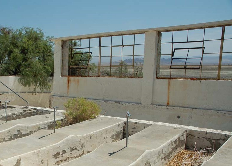 Taken from inside poolhouse ruins. A window frame stands without walls or a roof. In the foreground are empty bathtub-sized soaking pool depressions in the pavement.