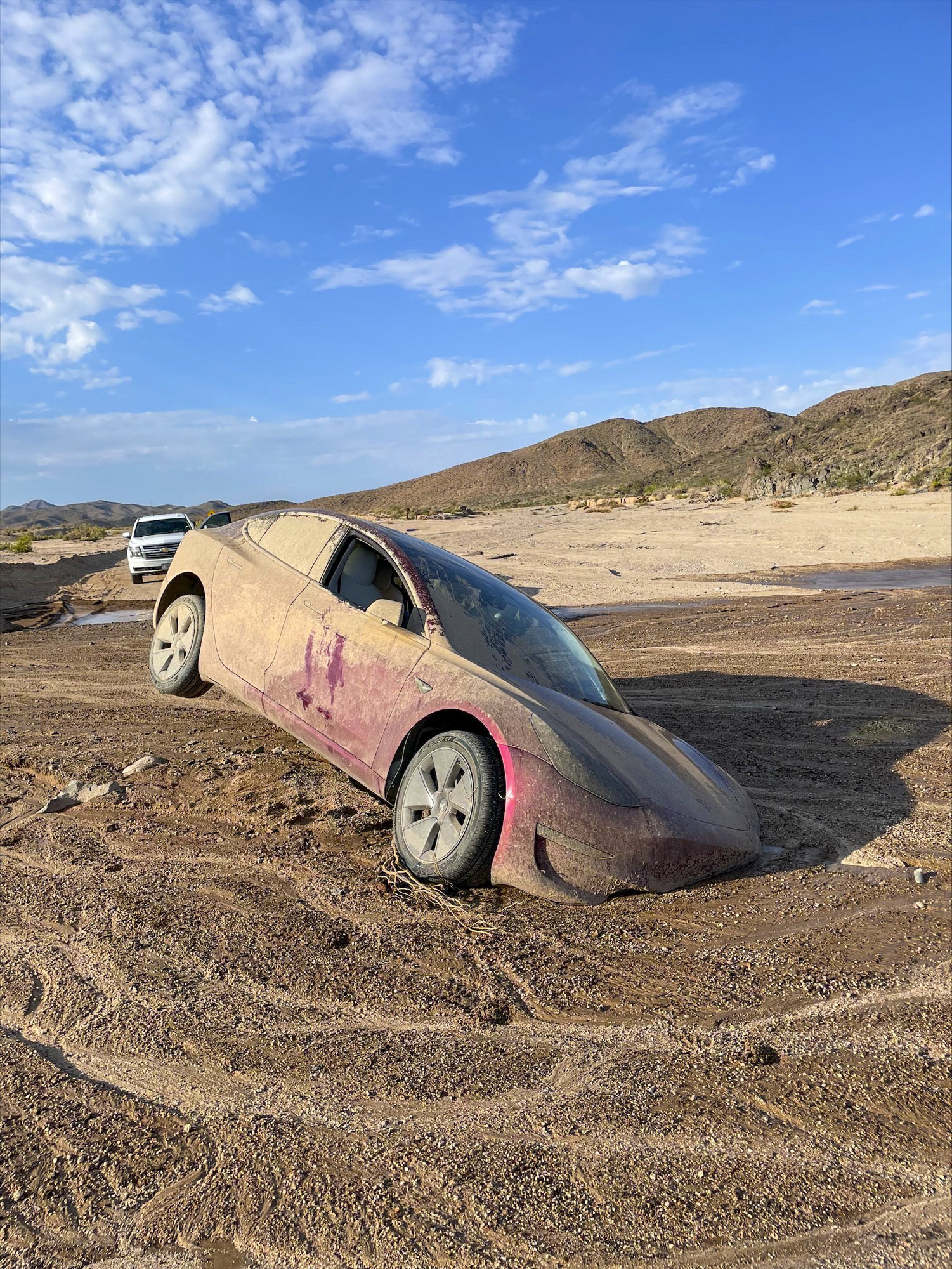 A red Tesla, almost completely covered in dried mud, stuck in a desert wash after recent monsoonal rains. The back passenger tire is in the air.