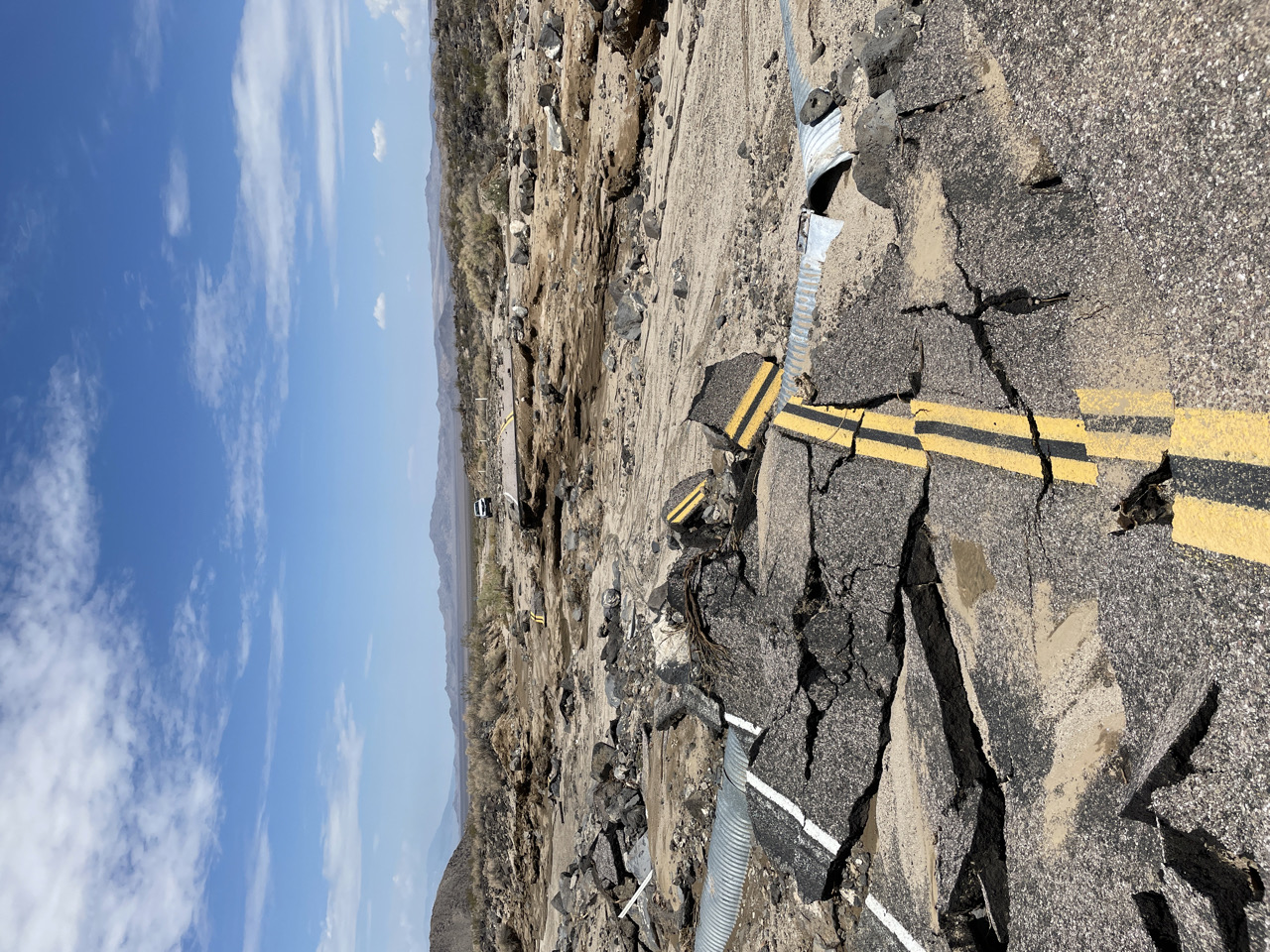 N Kelbaker - Seventeen Mile Point damage Road asphalt is crumbled up with a metal pipe sticking out. Clouds loom in the sky over the broken road.