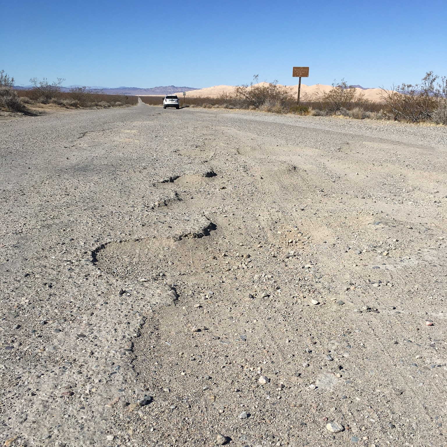 Pothole on dunes road A pothole with wavy irregular edges. In the background, the Kelso Dunes and a white vehicle.