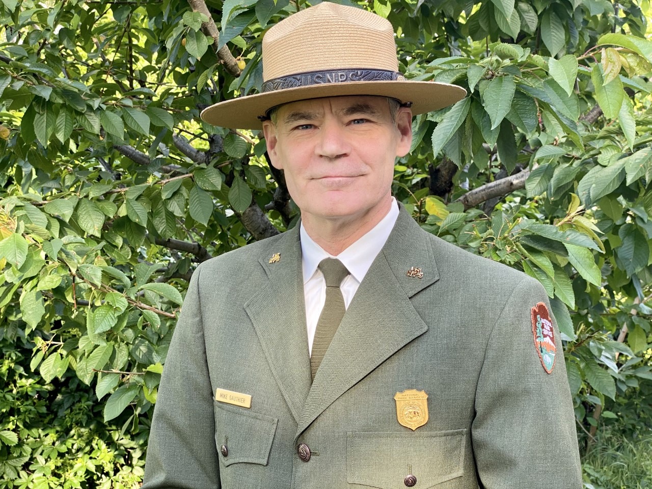 A man stands smiling in an NPS ranger dress uniform: flat top hat, long-sleeve green coat.