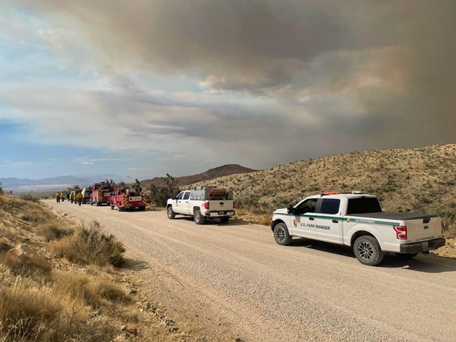 Fire trucks and other vehicles are parked along a gravel road with smoke in the background