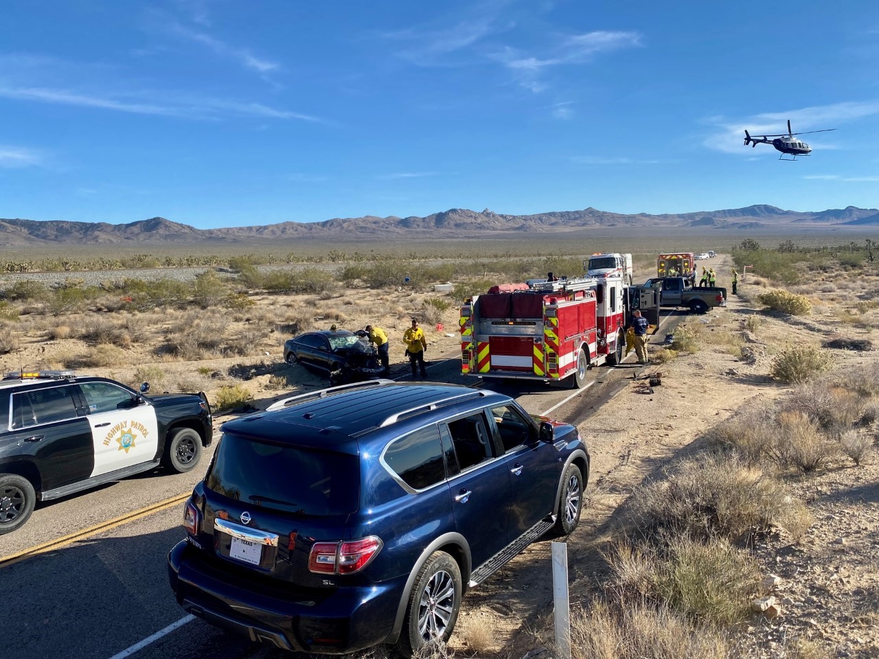 Emergency vehicles around a vehicle collision in the desert. a helicopter is leaving the scene.