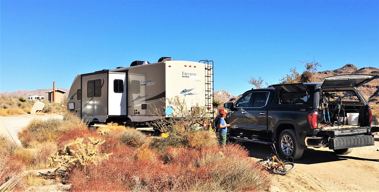 An RV trailer with slide out is pulled in to a campsite. A pickup truck and a young child with a bid face away is in the foreground