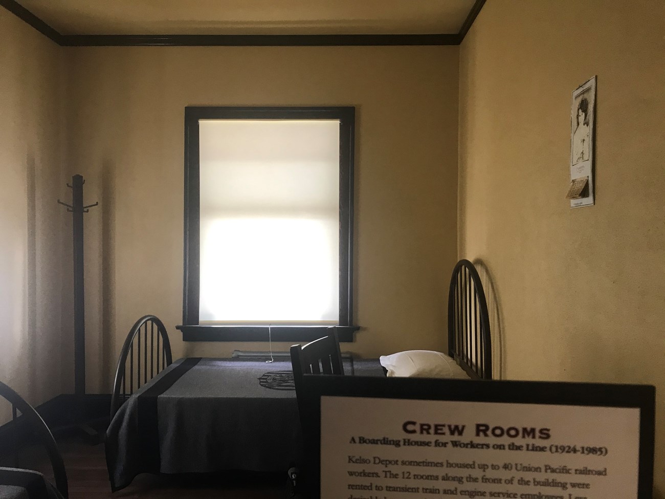 Crew bedrooms with window Kelso Depot.  A bed with a metal frame, blue Union Pacific sheets, and a white pillow.  An early 20th century calendar on the right wall. A coat stand in the left corner.  The edge of a second bed seen on the left.