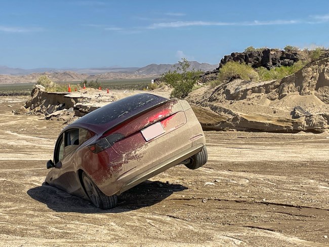 A red Tesla stuck in a desert wash after recent monsoonal rains. The back passenger tire is in the air