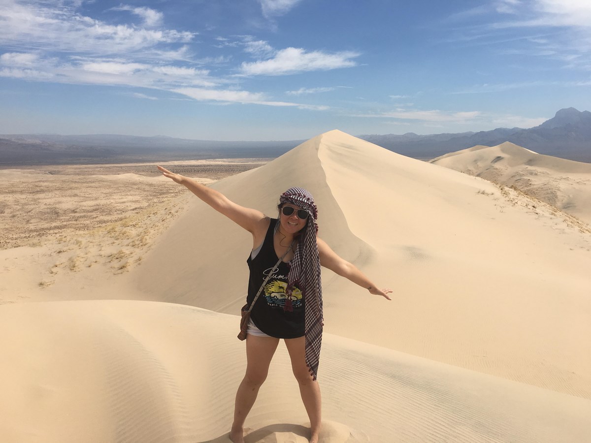 A young woman stands on top of a Kelso Sand Dune with arms outstretched.