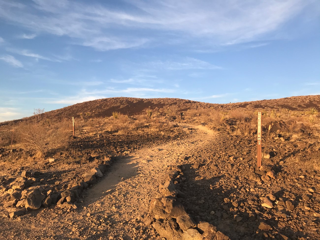 Sunset on the lava tube trail. Trail signs mark the junction to begin the final uphill hike to the lava tube. Lava and creosotes line the uphill trail.