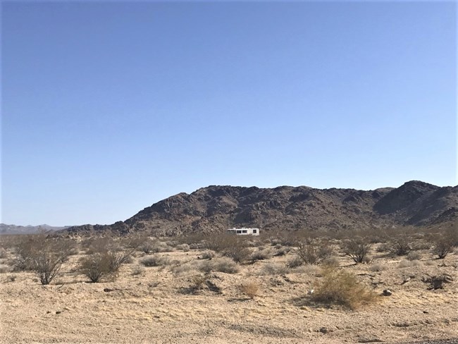 A RV camps at a undeveloped campsite surrounded by creosote. Mountains are in the background.
