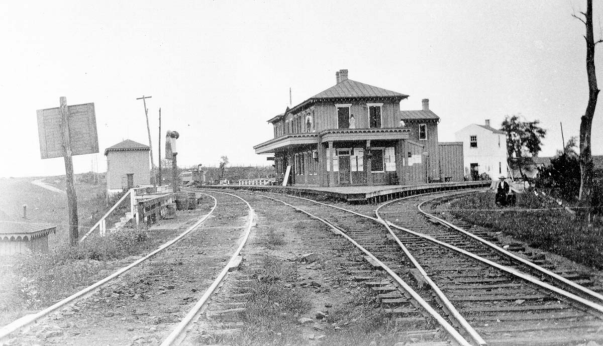 Black and white photo looking down railroad tracks at a two story train station.