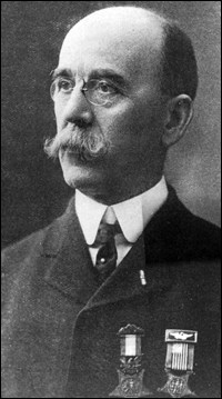 Older man with mustache wearing a suit with two medals pinned on chest.