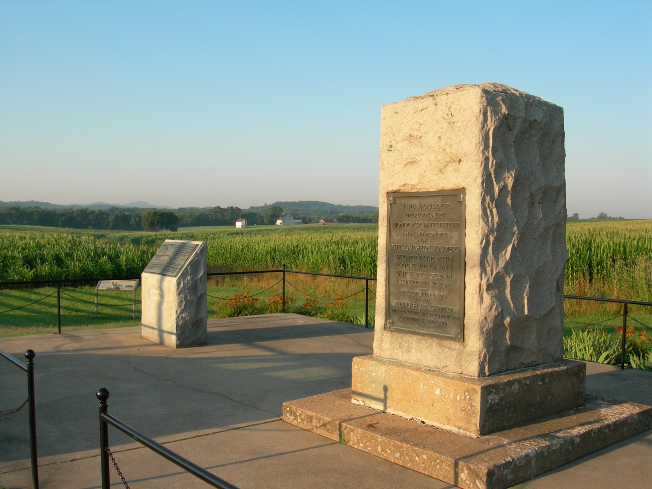 A rough cut 12 foot tall block of granite with a bronze plaque on the front sits in the front. Behind it is a smaller stone monument. Green farm fields are in the background.