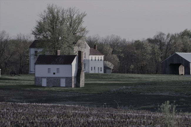 A two-story house made of stone on the first floor and wood on the second floor, with a larger all white house in background.