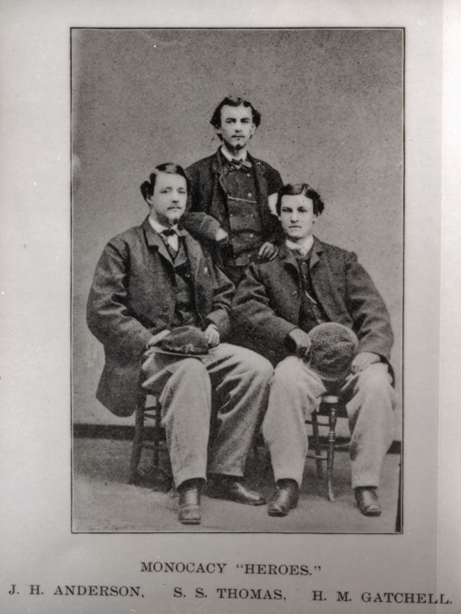 Three young men: two seated in front and third standing between them.
