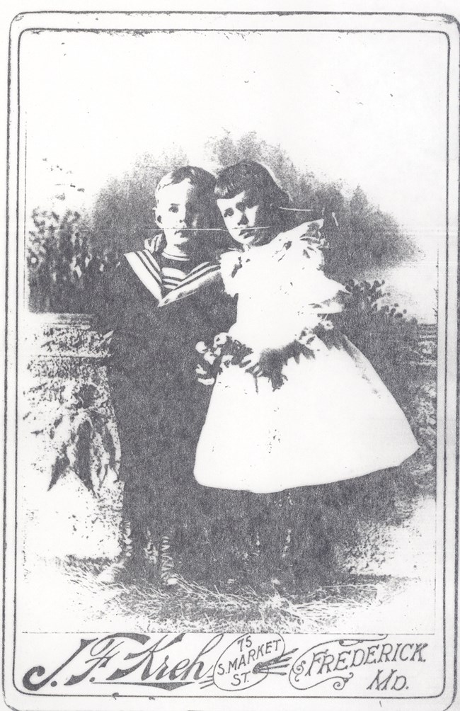 Two young children: on the left a boy wearing a dark sailor-style suit and on the right a girl in a white dress.