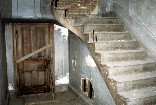 Decorative paint mimics wallpaper in a stairwell. There are holes in the walls and the railing is missing on the stairs.