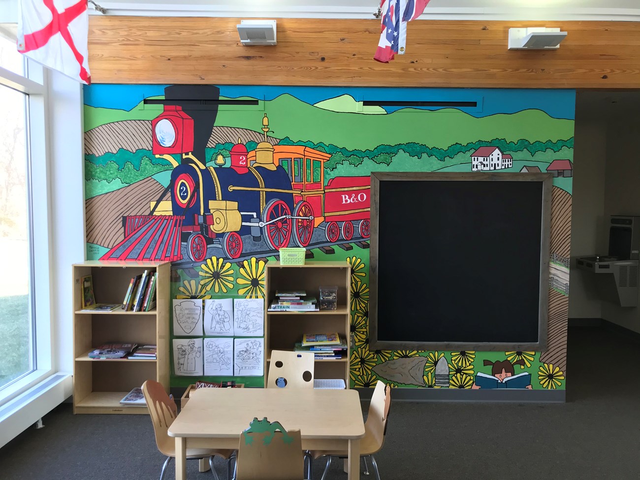 A cartoon of a 1830s locomotive with farm fields and a white house in the background decorates a wall. The mural also has black-eyed Susans, a projectile point, a minie ball, and a little girl reading in the lower right corner.