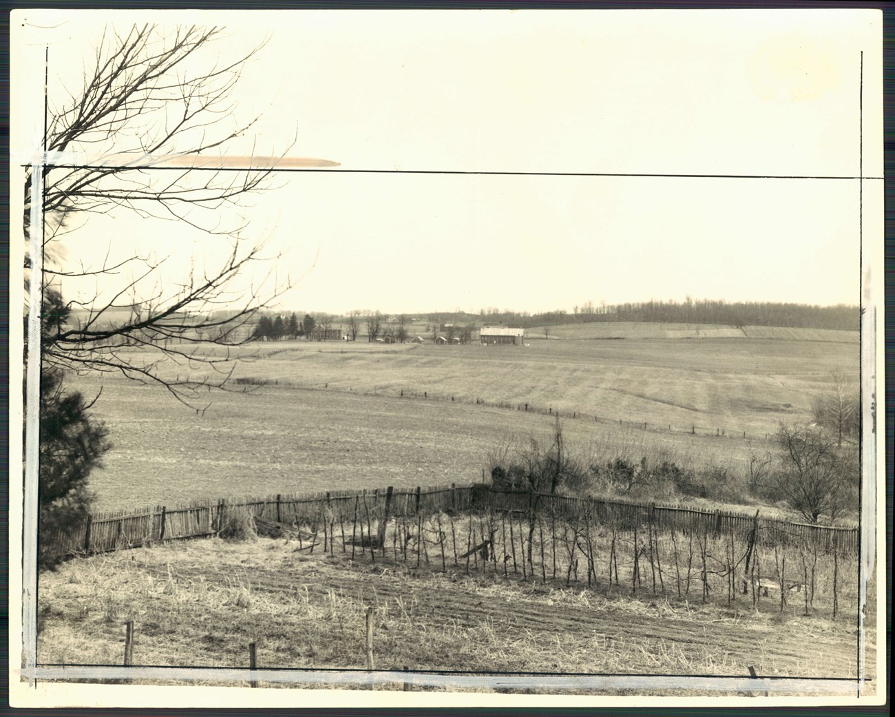 Black and white photo of open farm fields with a fenced garden in the foreground and a barn in the distance across empty fields.