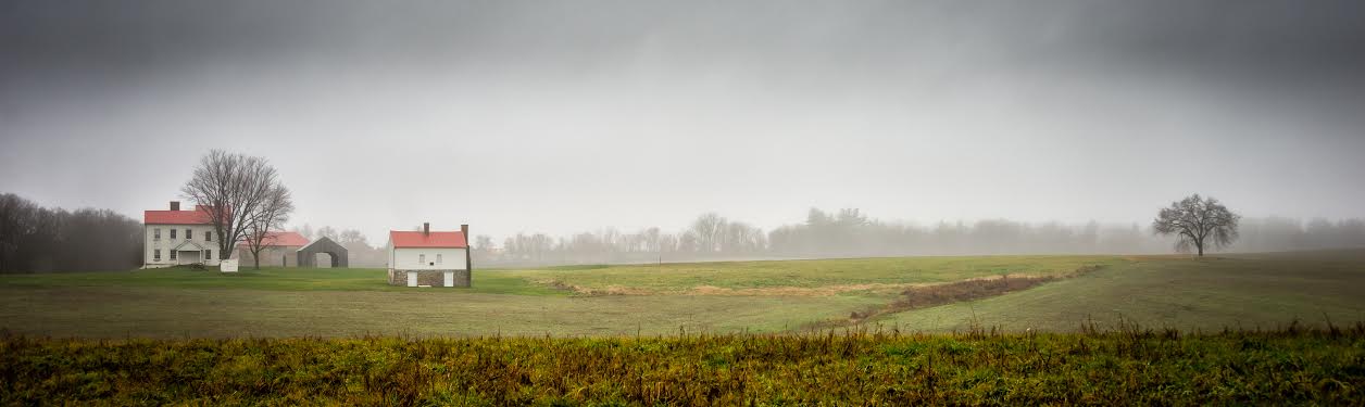 A white farm house and outbuildings with farm fields partially obscured by fog.
