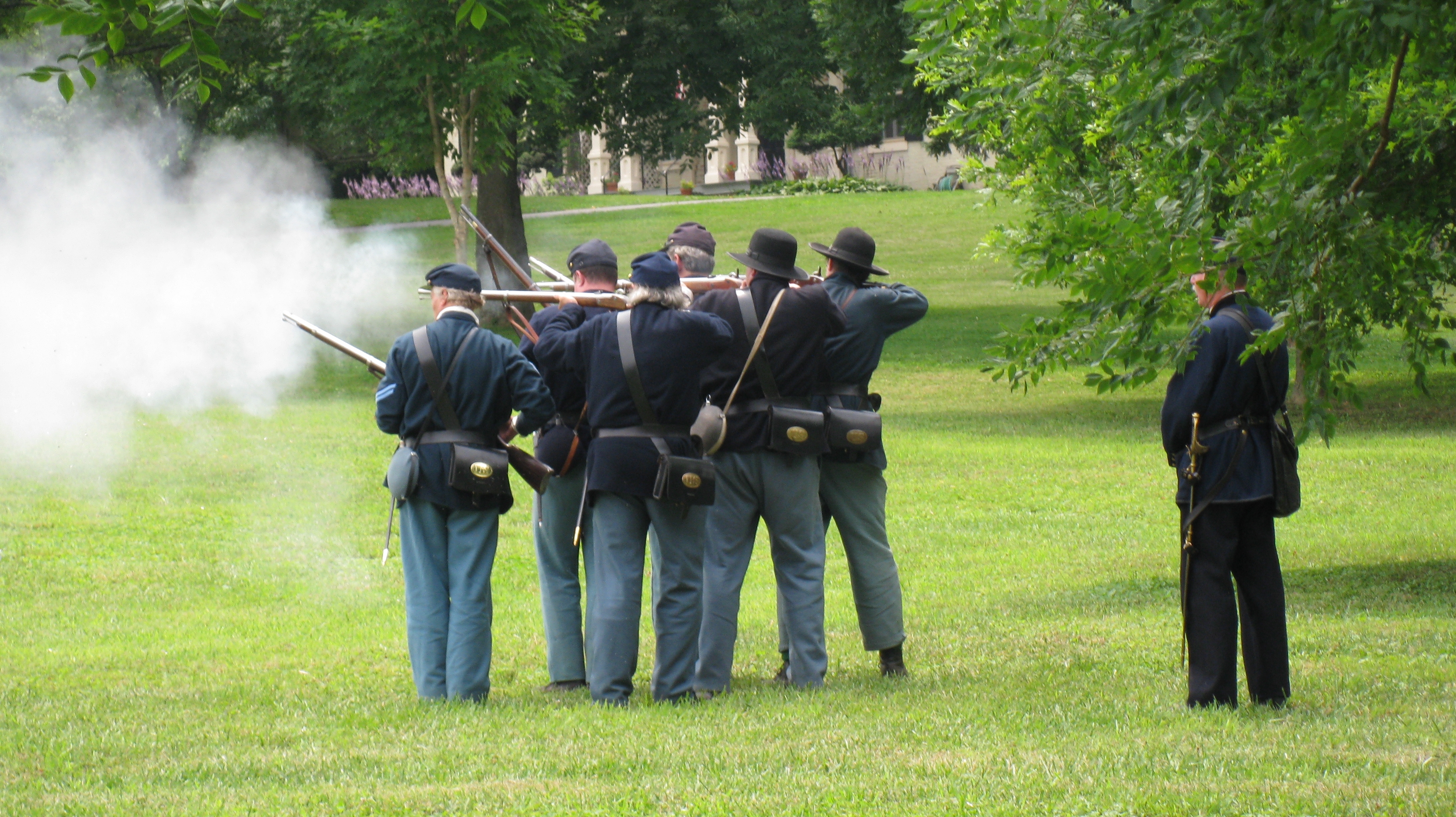 Six men in Civil War Union Army uniforms fire rifles while another man in uniform stands behind them.