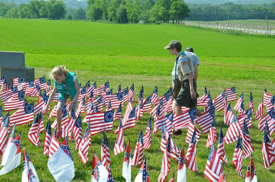 A boy in a scout uniform and a young girl set up historic flags on a lawn.