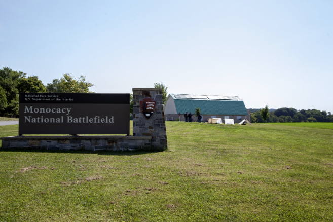 Entrance sign for Monocacy National Battlefield beside a grassy field, with the visitor center and tree-covered hills in the background