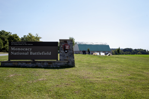 Entrance sign for Monocacy National Battlefield beside a grassy field, with the visitor center and tree-covered hills in the background
