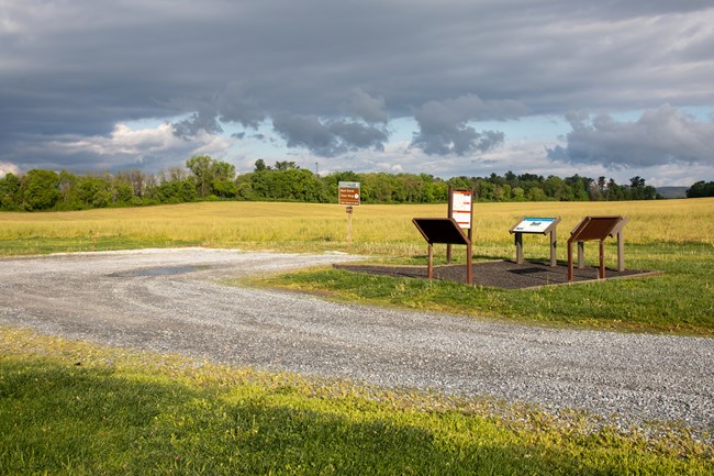 A gravel road leading to a gravel parking area with four information signs on a paved pad to the side of the parking area.