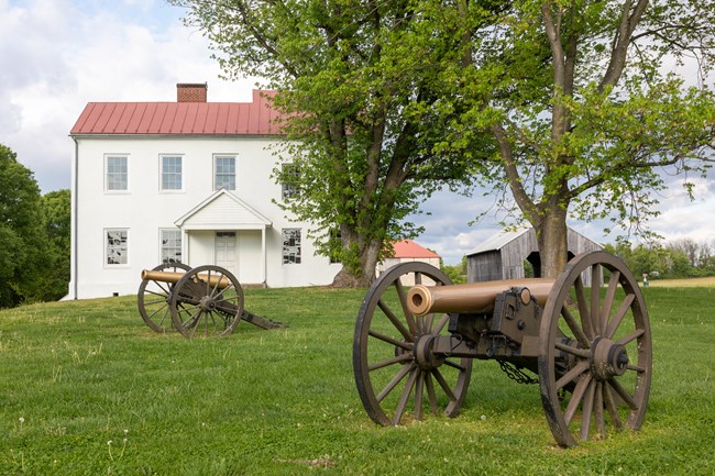 Two cannon on a grass field in front of a white two-store house with a red roof.