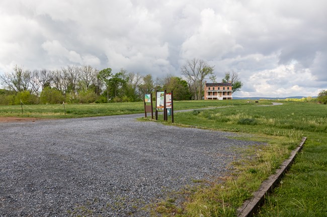 A gravel parking lot with three information signs and a gravel trail extending in the distance towards a two-story brick house surrounded by grassy fields.