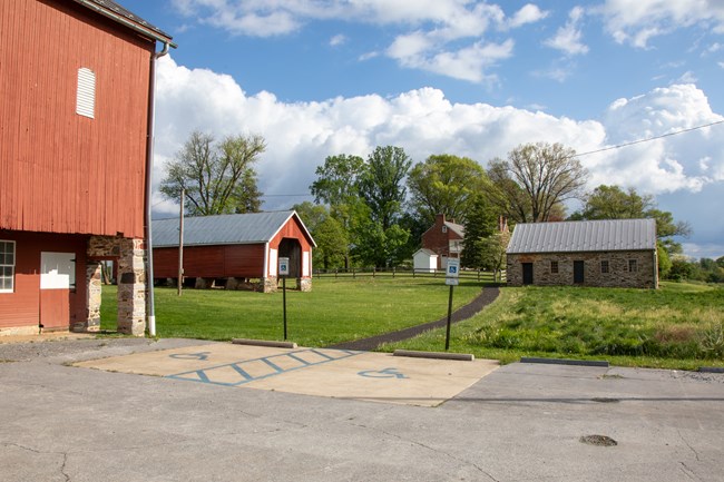 Two signed accessible parking spaces with an access aisle between them next to a red barn and a paved path leading toward a small stone building.