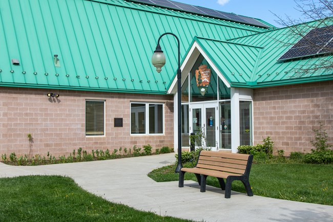 A sidewalk with a bench leads up to the glass double doors of a brick building with a turquoise metal roof.