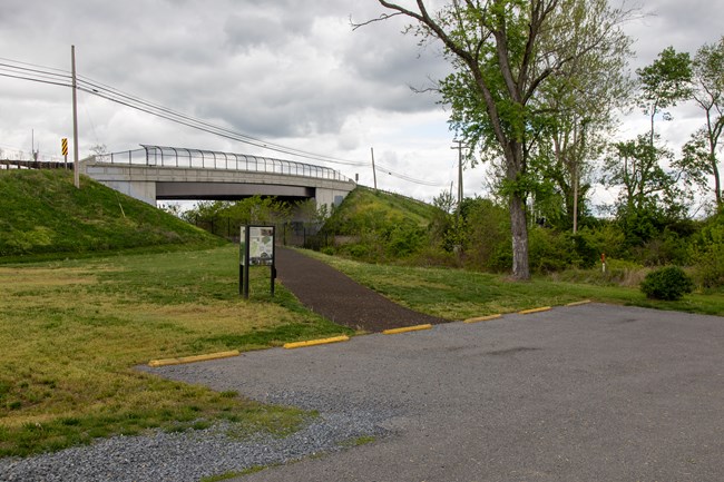 Five parking spaces next to a sign and a paved path leading under a road overpass.