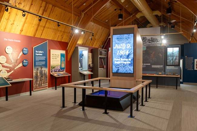 A high ceiling room with museum exhibits with photos and text and a large screen surrounded by a wood railing.
