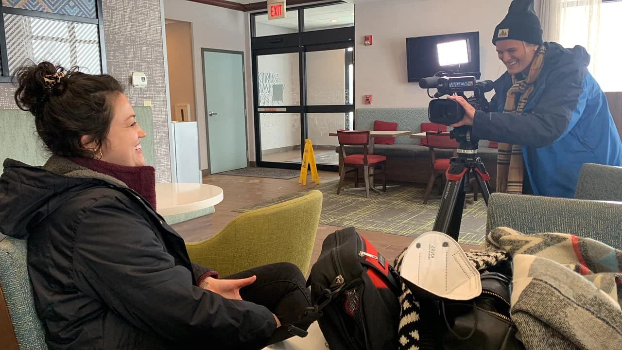 Setting is hotel lobby, entrance visible at rear. Seated woman wearing black coat in lower left corner, facing a camera and operator on the right.