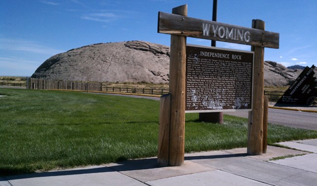A large wooden sign stands in front of a distant, giant rock.