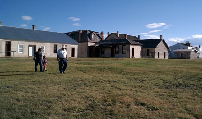 Two adults and one child walk across a grassy lawn in front of tan buildings with numerous chimneys. Small white clouds dot the sky. Two adults and one child walk across a grassy lawn in front of tan buildings with numerous chimneys. Small white clouds dot the sky.