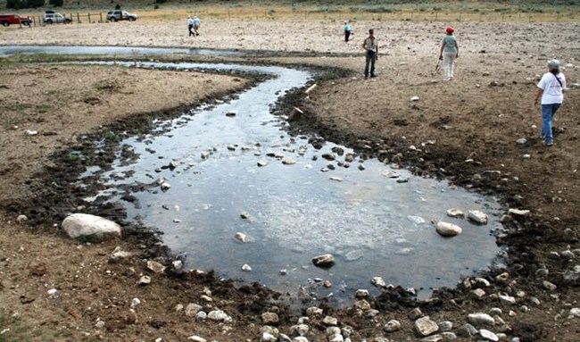People walk around a shallow pool of water surrounded by medium sized rocks. The water is in a teardrop shape. People walk around a shallow pool of water surrounded by medium sized rocks. The water is in a teardrop shape.