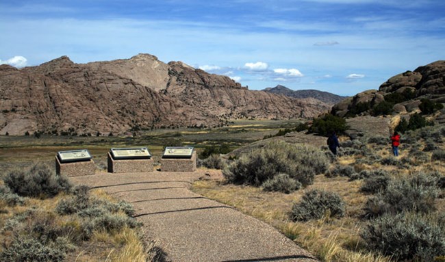 Two visitors walk on a trail in sagebrush near three wayside exhibits facing a large brown grey rock butte called Split Rock in Wyoming. Two visitors walk on a trail in sagebrush near three wayside exhibits facing a large brown grey rock butte called Split Rock in Wyoming.