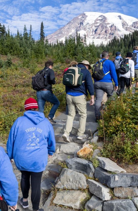 Group of people hikes up stone steps along a trail in a meadow with a glaciated mountain peak in the background.